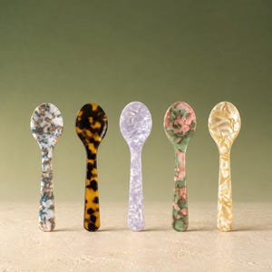 May include: Five small spoons arranged in a row, each featuring a distinct pattern. The spoons display a blue and white marble, a dark brown tortoiseshell, a solid light purple, a green and pink speckled, and a cream-colored pearlescent finish.