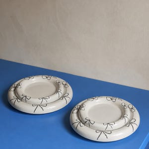 Puede incluir: Dos platos de cerámica blanca con un patrón de lazos negros dibujados a mano alrededor del borde elevado. Cada plato tiene un acabado liso y brillante, con un diseño circular que presenta un centro hundido y un borde ancho y curvo.