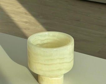 Puede incluir: Una taza de ónix verde claro con un cuerpo cilíndrico y una base corta y redondeada. La taza tiene una superficie lisa y pulida con vetas naturales. La taza está vacía y parece hecha a mano.