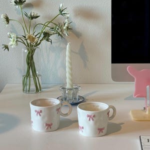 May include: Two white ceramic mugs decorated with pink bows are on a white surface. A clear glass vase with white flowers and a twisted white candle in a glass holder are also present. The scene is bright and airy.