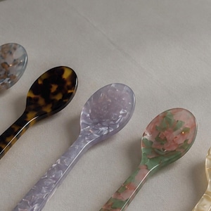May include: A collection of five small spoons made from polished resin with various patterns. The spoons feature a tortoiseshell design, a light lavender marbled finish, a pink and green floral-like pattern, and a cream-colored marbled texture.