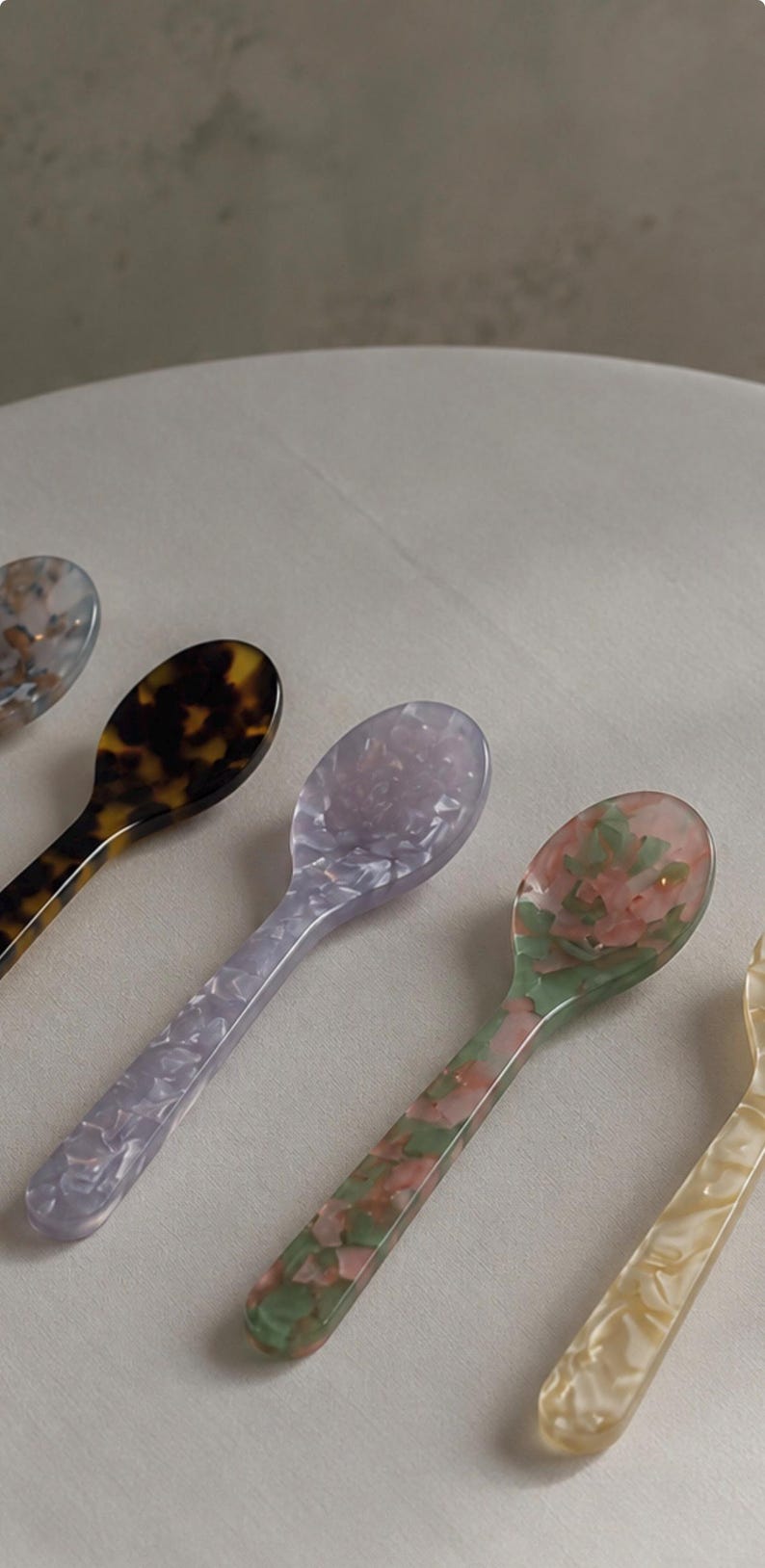 May include: A collection of five small spoons made from polished resin with various patterns. The spoons feature a tortoiseshell design, a light lavender marbled finish, a pink and green floral-like pattern, and a cream-colored marbled texture.