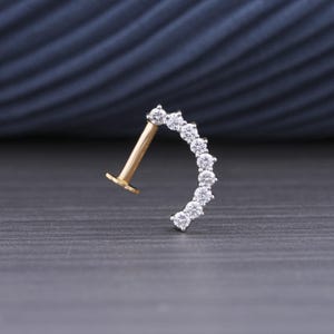 May include: A gold-colored, curved piercing stud with a row of seven clear, round gemstones. The post and base of the stud are gold, while the gemstones are set in a silver-colored metal. The jewelry is set against a dark gray background.