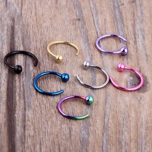 May include: A collection of nose rings in various colors, including black, gold, blue, silver, purple, pink, and iridescent. Each ring features a small ball at the end. The jewelry is displayed on a wooden surface.