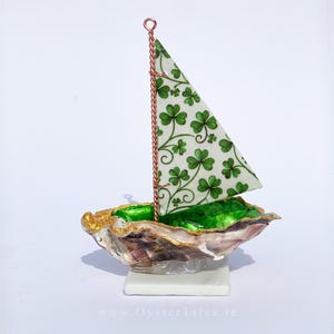 Peut inclure: Un voilier décoratif en coquille d'huître, avec un intérieur vert et doré. La voile est blanche avec un motif de trèfles verts, et le mât est en fil de cuivre. Le bateau repose sur une petite base blanche.