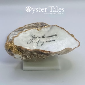 May include: An oyster shell with a gold-painted edge and the text "You're the measure of my dreams" in black script. The shell is presented on a small, white rectangular base. The Oyster Tales logo is visible in the background.