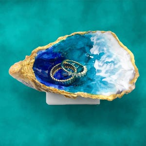 May include: An oyster shell jewellery dish with a gold rim, filled with blue and white resin, holding two gold rings. The dish sits on a white base against a teal background. The rings have clear stones.
