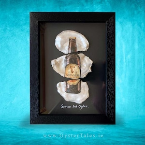 Könnte beinhalten: Gerahmtes Kunstwerk mit einer Guinness Extra Stout-Flasche zwischen drei Austernschalen auf schwarzem Hintergrund. Der Text "Guinness And Oysters" steht darunter. Der Rahmen ist schwarz.
