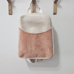 Puede incluir: Una mochila cuadrada con la parte superior color crema y un bolsillo delantero color melocotón cuelga de un gancho de madera. La bolsa tiene un asa y correas de color crema. Tres ganchos de madera están montados en una pared blanca.