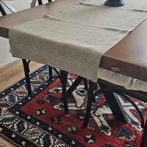 May include: A dining table with a light-coloured runner and black metal legs, set on a red and blue patterned rug. Wooden chairs are in the background, along with a decorative lantern. The table is made of wood.