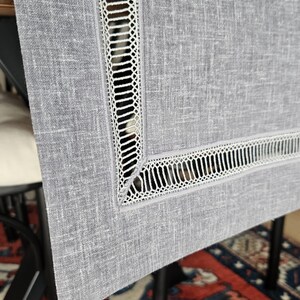 May include: A close-up of a grey linen table runner with a white, openwork lace border. The runner is draped over a table, showing the texture of the fabric and the intricate design of the lace. The runner adds a touch of elegance.