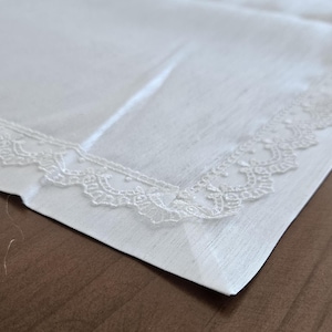 Puede incluir: Un pañuelo blanco cuadrado con un delicado borde de encaje festoneado. La tela parece ser un material suave y tejido. El ribete de encaje añade un toque de elegancia al diseño clásico.