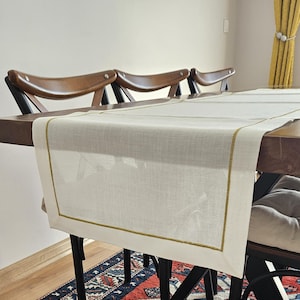 May include: A cream linen table runner with a gold border is laid on a dark wooden table. The runner is rectangular. Three wooden chairs are positioned behind the table. A patterned rug is visible beneath the table. The table runner measures approximately 180 cm in length.