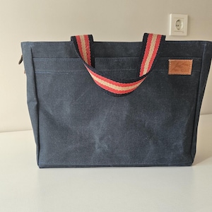 Puede incluir: Un bolso tote azul marino con un asa a rayas en rojo, beige y blanco. El bolso tiene un pequeño parche de cuero marrón en la parte delantera. El bolso es rectangular y parece estar hecho de lona.