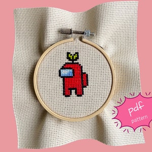 Könnte beinhalten: Kreuzstich-Stickerei eines roten Among Us-Charakters mit einer grünen Krone, in einem Holzreifen. Das Design befindet sich auf beigem Stoff. Der Text "pdf pattern" steht in einer rosa Sprechblase.