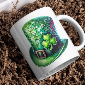 以下が含まれることがあります： 緑、青、銀のラメで飾られたレプリコーンの帽子が描かれた白いセラミック製のマグカップ。帽子には、シャムロックが乗っており、黒いバンドに金色のバックルが付いています。