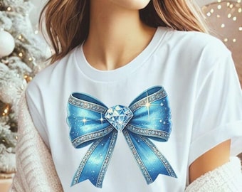 May include: White t-shirt featuring a large, detailed blue bow with a sparkling diamond center. The bow has glitter accents and a ribbon design. The shirt is a classic crew neck style.