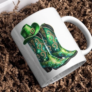 以下が含まれることがあります： 緑と金の色のカウボーイブーツと緑の帽子が描かれた白いセラミックマグカップ。ブーツはスパンコールで覆われています。
