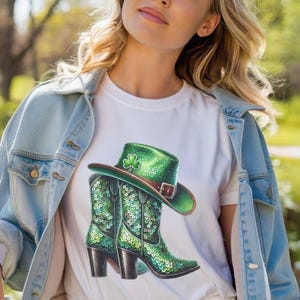 以下が含まれることがあります： 緑のキラキラしたカウボーイブーツと帽子がデザインされた白いTシャツ。帽子にはクローバーが飾られています。