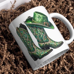 以下が含まれることがあります： 緑と金のデザインが施された白いセラミック製のマグカップ。カウボーイハットとブーツ2つが描かれています。ハットとブーツは、スパンコールで覆われています。