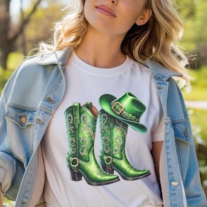 以下が含まれることがあります： 緑色のカウボーイハットと緑色のカウボーイブーツ2つが描かれた白いTシャツ。ブーツは緑と金のデザインで、ラインストーンが飾られています。