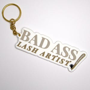 May include: White keychain with gold lettering that reads "Bad Ass Lash Artist" with a small black and white lash brush graphic and the text "Un-Stripped" on the right side.