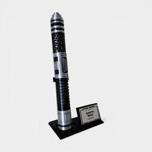 Puede incluir: Una r&eacute;plica del sable de luz Darth Maul de Star Wars. El sable de luz es negro y plateado con un dise&ntilde;o detallado. Se muestra en un soporte negro con una placa con el texto "STAR WARS Darth Maul - Lightsaber."
