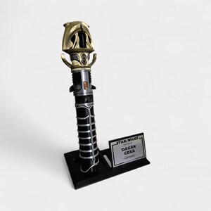 May include: A black and gold lightsaber with a silver accent on the hilt. The lightsaber is on a black stand with a silver plaque that reads "Star Wars: The Bad Batch Dagan Gera Lightsaber".