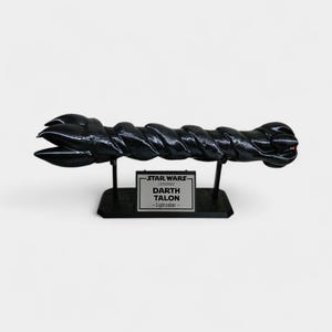 May include: A black Darth Talon Lightsaber replica from Star Wars Legends, displayed on a black stand. The lightsaber has a twisted design and a red accent at the end. The stand features a silver plaque with the text "STAR WARS LEGENDS DARTH TALON - Lightsaber".