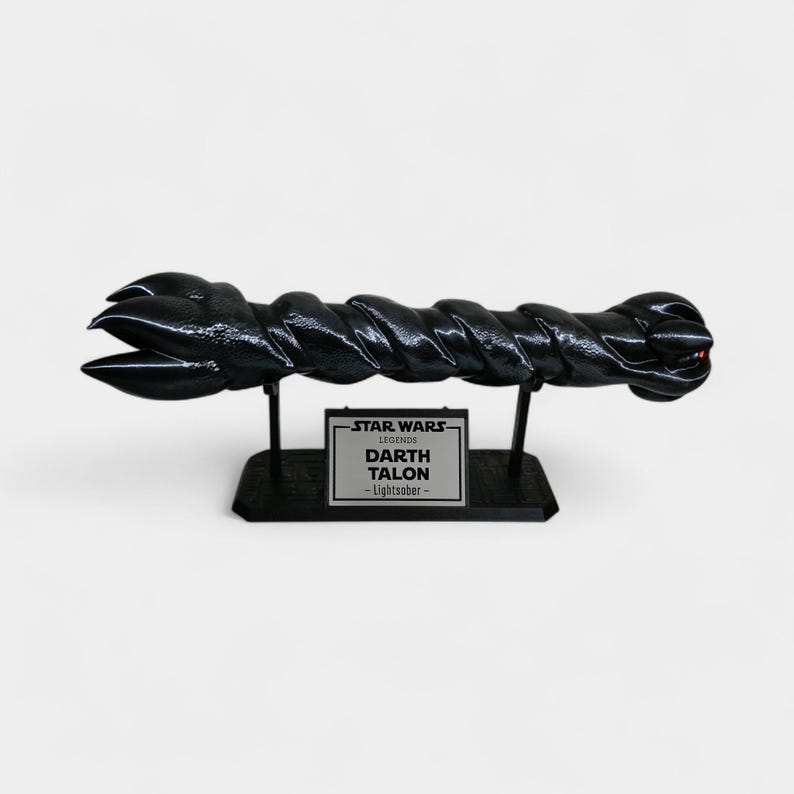 May include: A black Darth Talon Lightsaber replica from Star Wars Legends, displayed on a black stand. The lightsaber has a twisted design and a red accent at the end. The stand features a silver plaque with the text "STAR WARS LEGENDS DARTH TALON - Lightsaber".