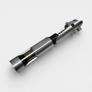 May include: A silver-colored lightsaber prop with a detailed design. The prop has a cylindrical body with a black grip and a partially exposed internal structure. The end features a series of metallic components and a small, circular button.