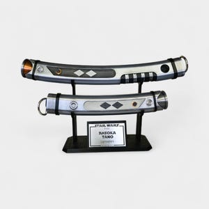 Peut inclure: Deux poign&eacute;es de sabre laser argent&eacute;es et incurv&eacute;es avec des accents noirs et des incrustations en forme de losange, sur un support noir. Une plaque indique : STAR WARS, AHSOKA TANO, Lightsabers. Les poign&eacute;es ont des finitions m&eacute;talliques.