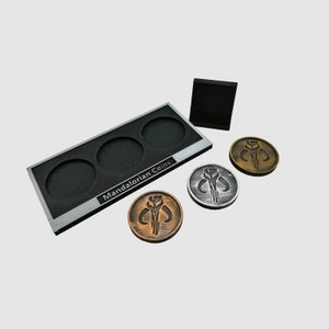 May include: A set of three Mandalorian coins, two silver and one bronze, with a black display stand. The stand has the words "Mandalorian Coins" printed on it.
