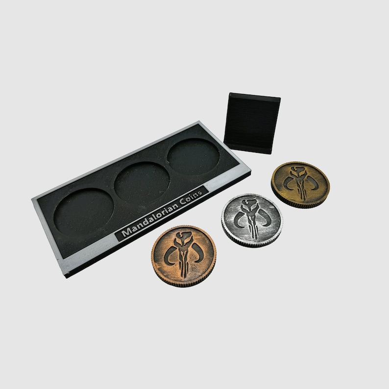May include: A set of three Mandalorian coins, two silver and one bronze, with a black display stand. The stand has the words "Mandalorian Coins" printed on it.