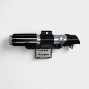 May include: A replica of Darth Vader's lightsaber from Star Wars. The lightsaber is black and silver with a rectangular base that reads "Star Wars Darth Vader Lightsaber". The lightsaber is mounted on a white background.