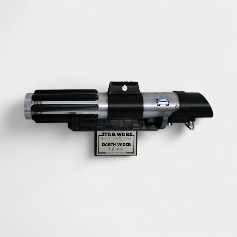 May include: A replica of Darth Vader's lightsaber from Star Wars. The lightsaber is black and silver with a rectangular base that reads "Star Wars Darth Vader Lightsaber". The lightsaber is mounted on a white background.