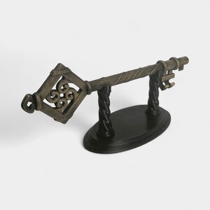 May include: A decorative bronze-colored key on a black stand. The key has an ornate, openwork design on the head and a twisted shaft. The stand is oval-shaped with two twisted supports.