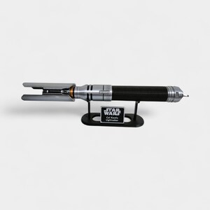 May include: A silver and black lightsaber with a textured grip, displayed on a black stand. The lightsaber has a distinctive blade emitter design. The stand features a Star Wars logo and the text "Cal Kestis Lightsaber."