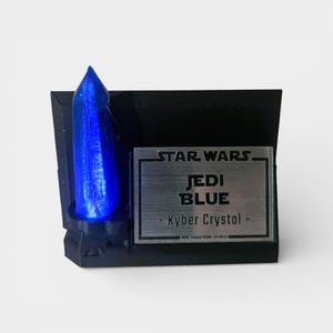 May include: A black display stand featuring a blue Kyber crystal. The stand has a silver plaque with the words "STAR WARS JEDI BLUE - Kyber Crystal". The crystal is a vibrant blue color and is the focal point of the display.