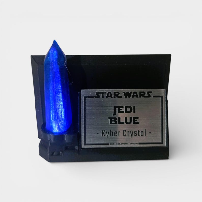 May include: A black display stand featuring a blue Kyber crystal. The stand has a silver plaque with the words "STAR WARS JEDI BLUE - Kyber Crystal". The crystal is a vibrant blue color and is the focal point of the display.