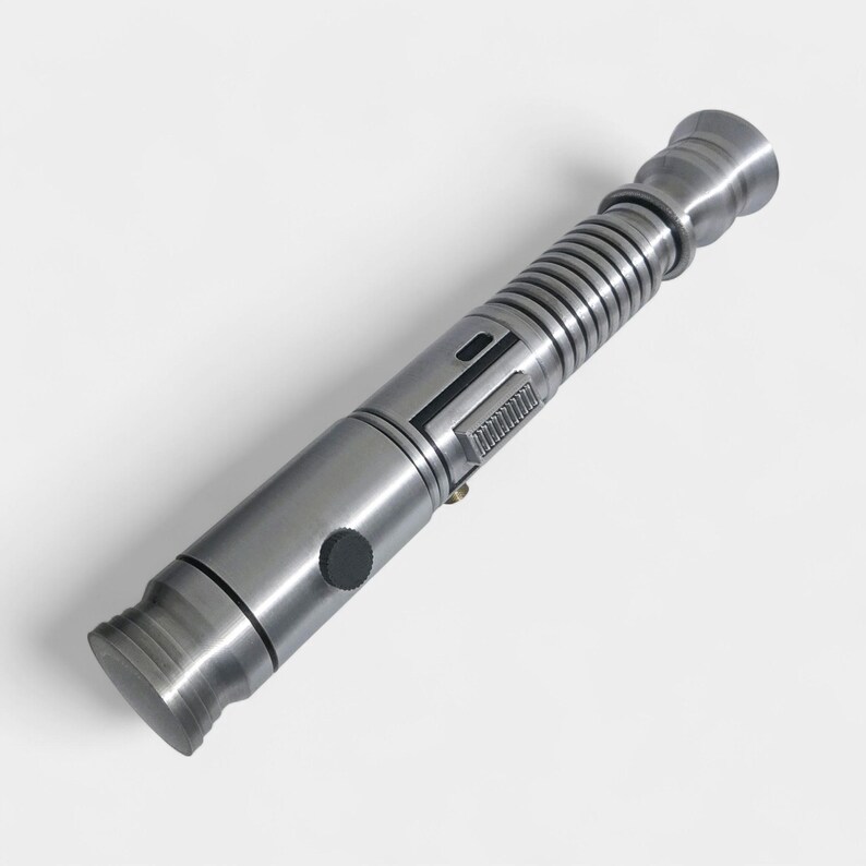 May include: A metallic silver lightsaber prop with detailed ridges and a black button. The prop has a cylindrical design with a textured grip and a flared emitter. The overall aesthetic is futuristic and industrial.