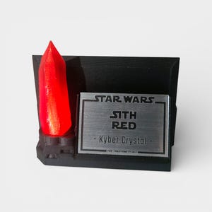 May include: A black display stand featuring a vibrant red Kyber crystal. The stand includes a metallic plaque with the text "STAR WARS SITH RED - Kyber Crystal." The base is rectangular, and the crystal is prominently displayed.