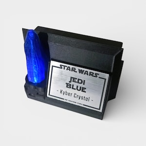 May include: A black display stand featuring a glowing blue Kyber Crystal. The stand has a silver plaque with the words "STAR WARS JEDI BLUE - Kyber Crystal". The design is a collectible item.