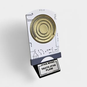 May include: A white and gray 3D printed model of the Death Star plans from Star Wars. The plans are on a stand with a black base and a gray top.