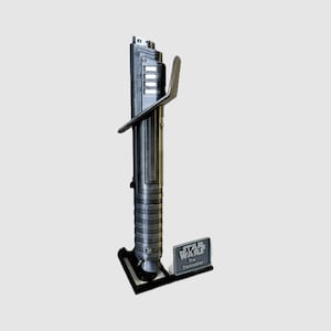 Op de afbeelding: Een zwarte lightsaber-prop met een zilveren handvat en een zwarte standaard. De standaard heeft een klein plakkaat met de tekst "Star Wars The Dark Saber".