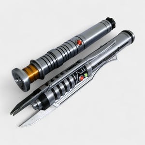 May include: Two metallic lightsabers with silver and black details. One has a red button and a gold-colored base. The other has a red and green button and a blade-like design. The lightsabers are on a white background.
