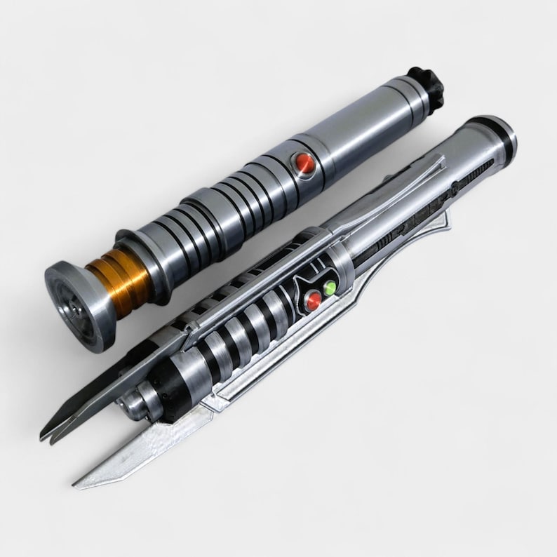 May include: Two metallic lightsabers with silver and black details. One has a red button and a gold-colored base. The other has a red and green button and a blade-like design. The lightsabers are on a white background.