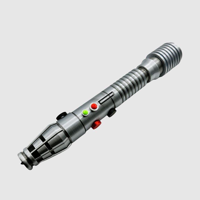 May include: A silver lightsaber with a black handle and a green and red light on the side. The lightsaber has a ribbed design and a black tip.