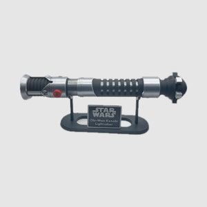 May include: A silver and black lightsaber replica on a black stand with a silver plaque that reads "STAR WARS Obi-Wan Kenobi Lightsaber".