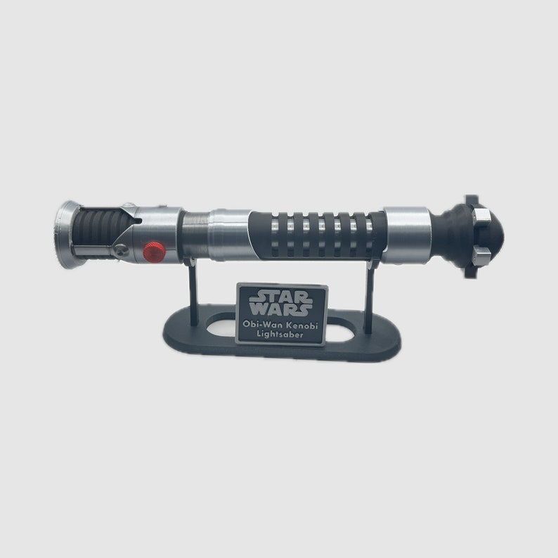 May include: A silver and black lightsaber replica on a black stand with a silver plaque that reads "STAR WARS Obi-Wan Kenobi Lightsaber".